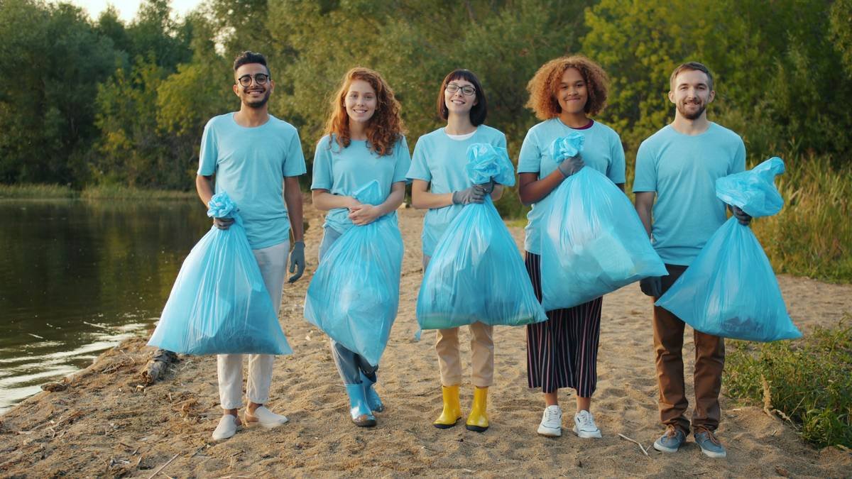 Portrait of multi-ethnic group of volunteers with bin bags on clean lake shore, men and women are standing together smiling looking at camera. Youth and charity work concept.